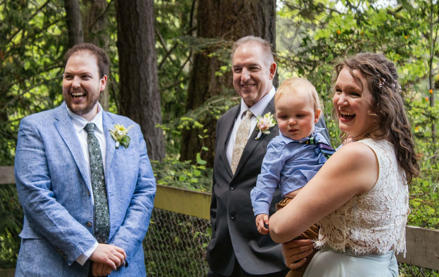 A joyful outdoor wedding moment captured in a wooded setting with people in formal attire and a small child.