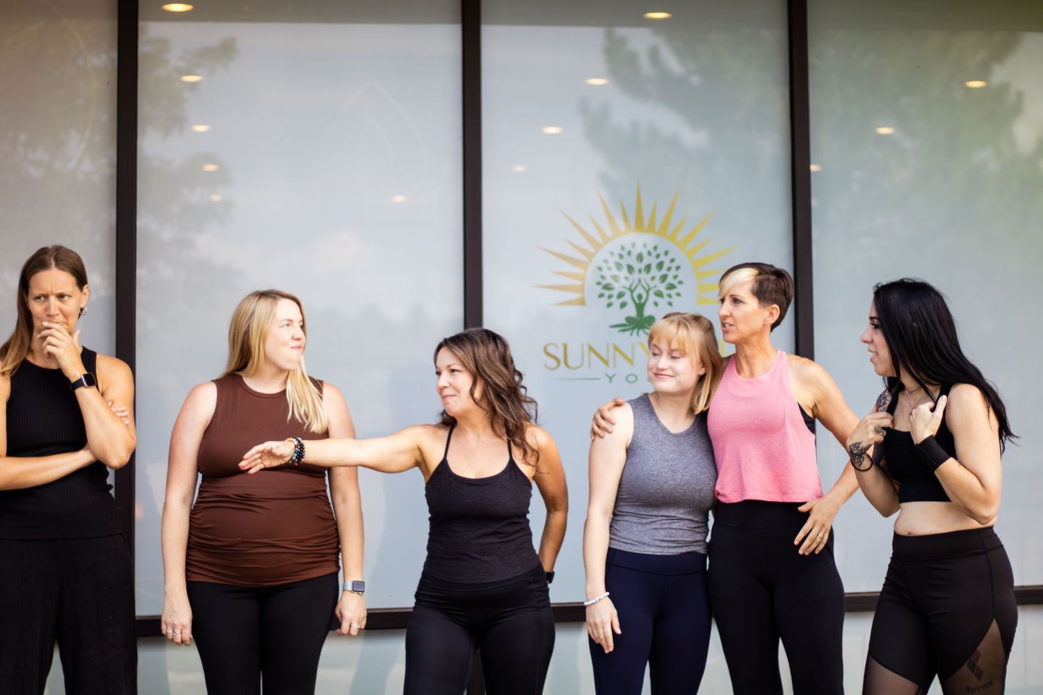 A group of women in workout attire stand in a line against a white wall while conversing and socializing.