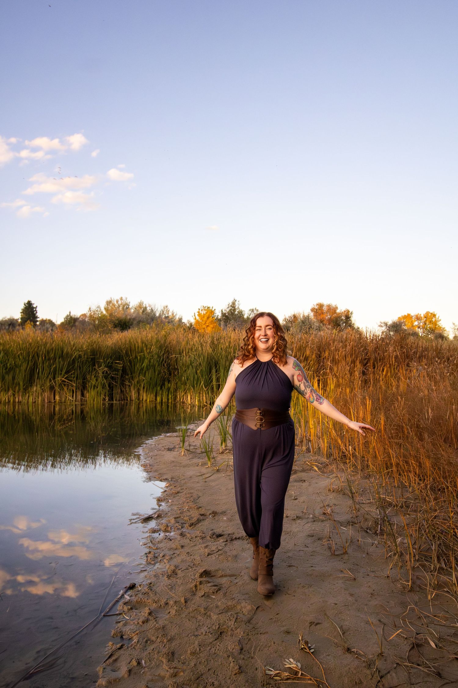 A person walks along a sandy shoreline path with tall reeds and water at sunset, creating a serene autumn scene.