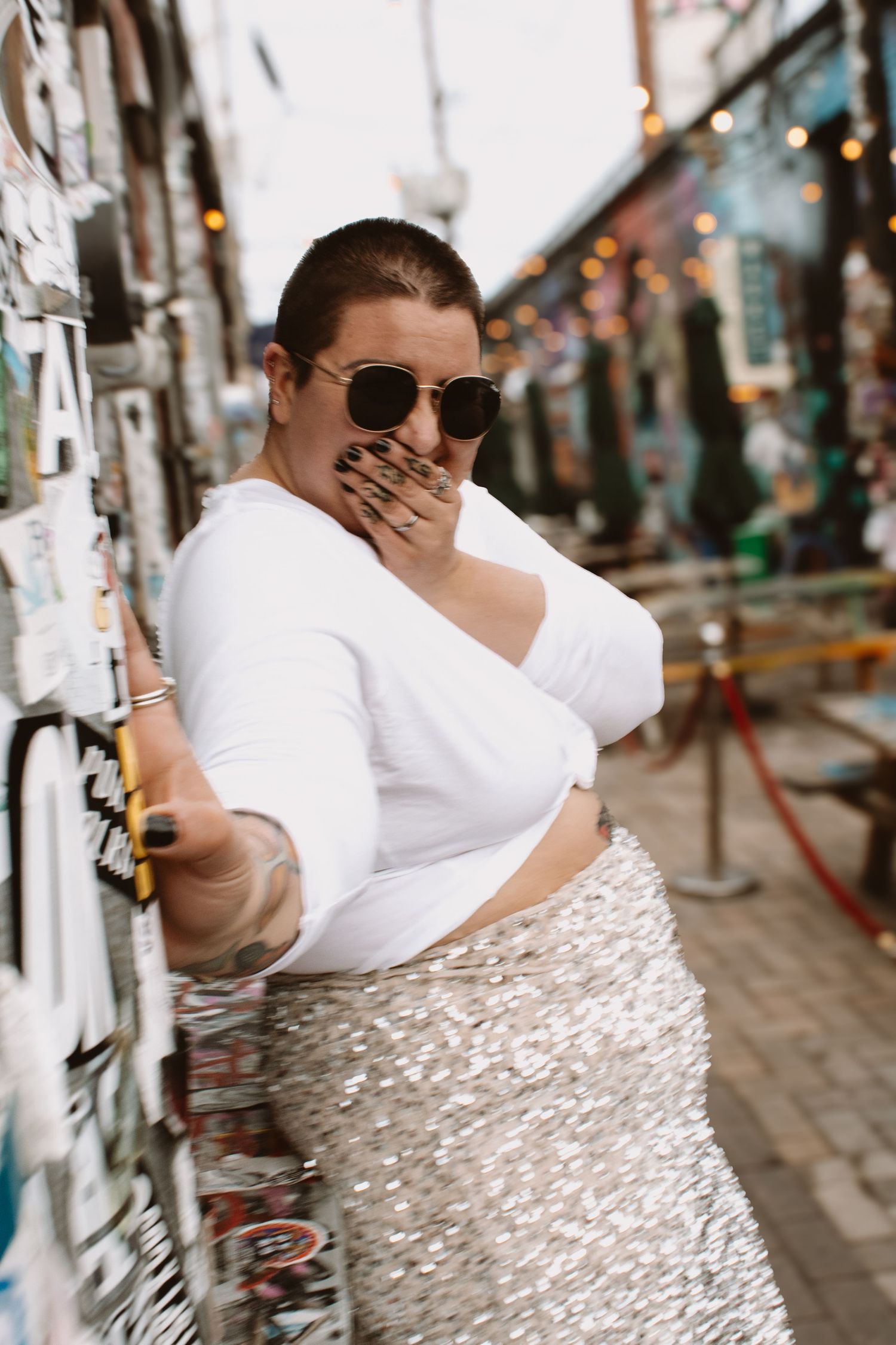 A person in a white top and patterned skirt poses against a textured wall in an urban setting.