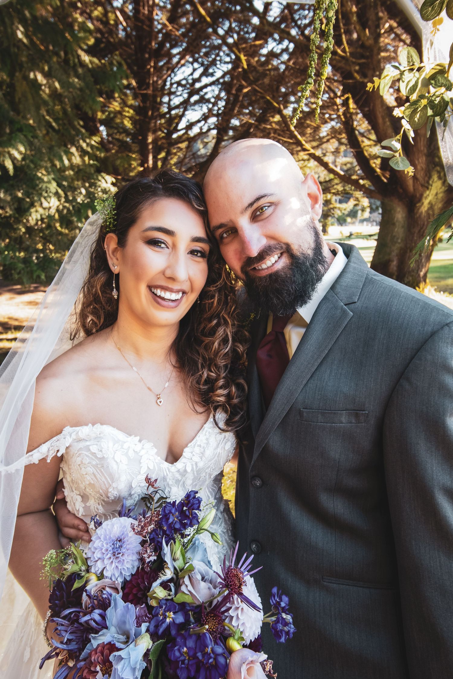 A happy couple poses outdoors on their wedding day with a stunning purple and blue floral bouquet.