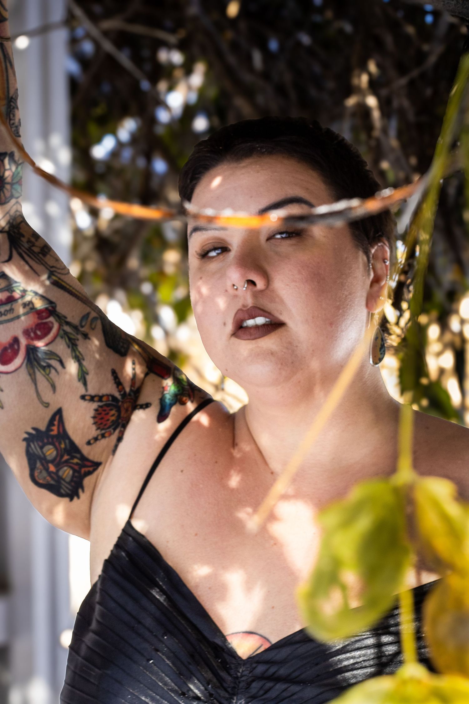 A portrait in dappled sunlight showing a dark-haired figure in black with visible arm tattoos among tree foliage.