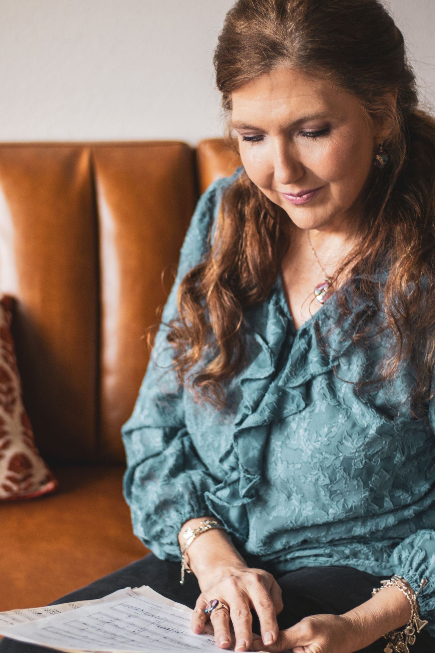 Someone wearing a teal ruffled blouse sits on a brown leather couch while reading documents.