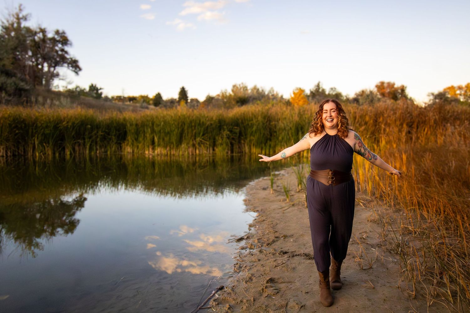 A person walks along a tranquil lakeside path at sunset with golden light illuminating the reeds and water.