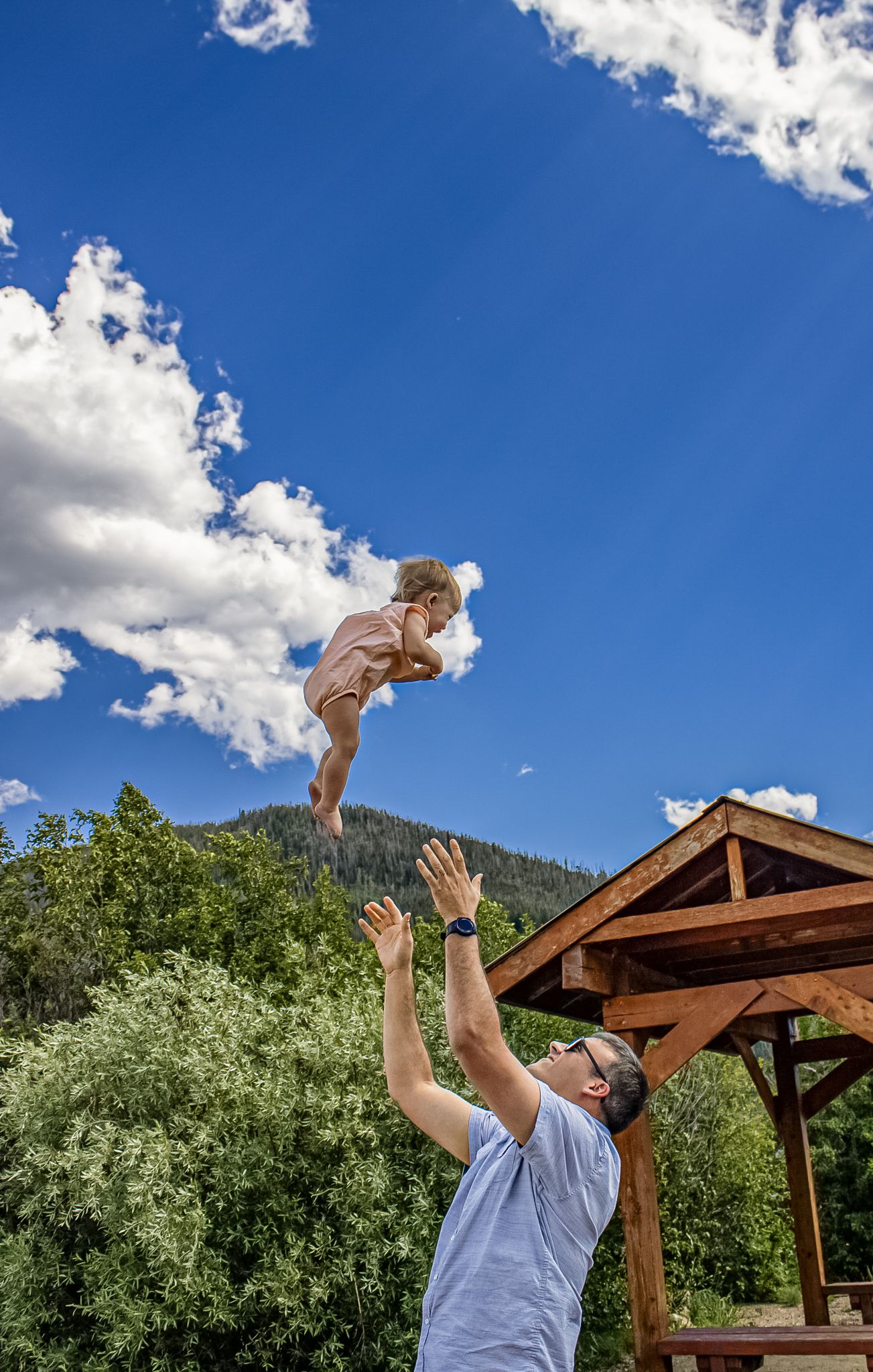 Parent and child playing together outdoors under blue sky and white clouds near wooden gazebo structure.