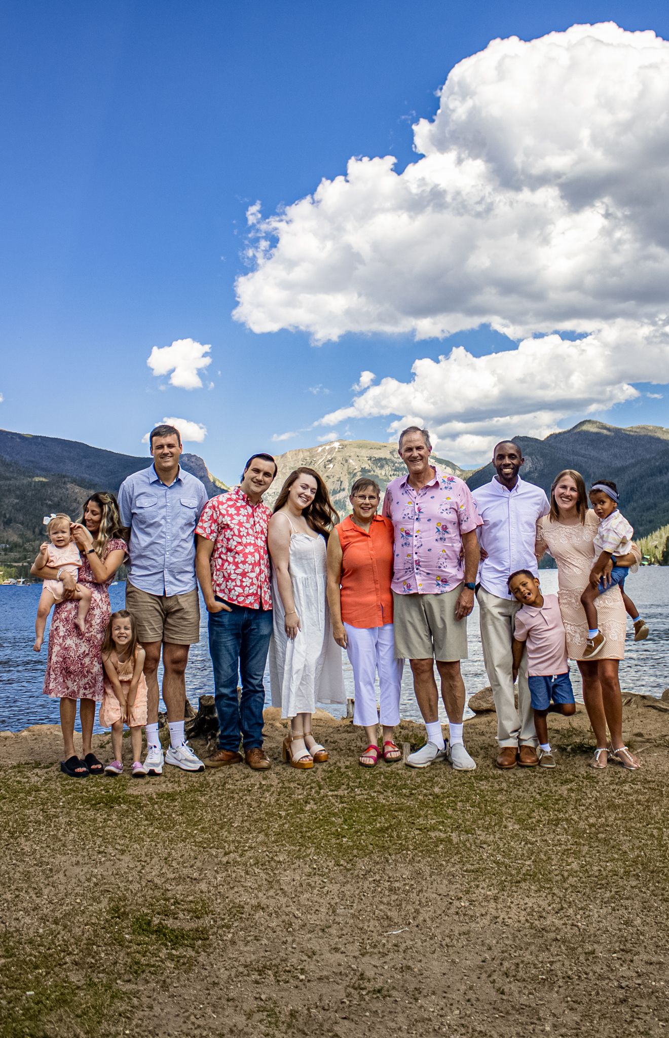 A group of people wearing coordinated pink and white outfits pose together on a grassy hill with mountains and lake in background.