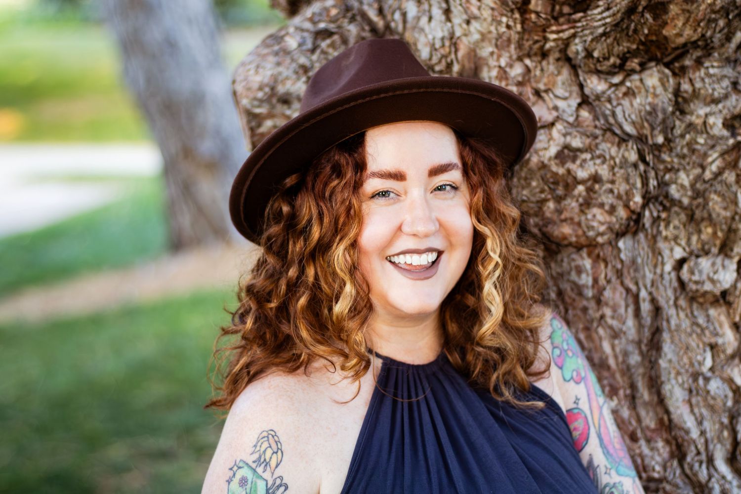 A woman with curly hair and a hat smiles while leaning against a tree trunk in an outdoor setting.