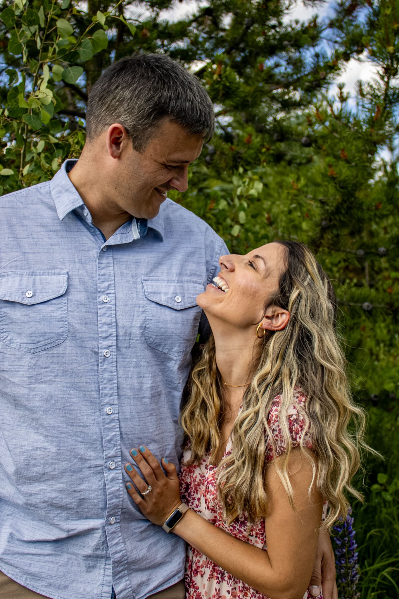 A couple shares a tender moment outdoors surrounded by lush greenery and trees on a sunny day.