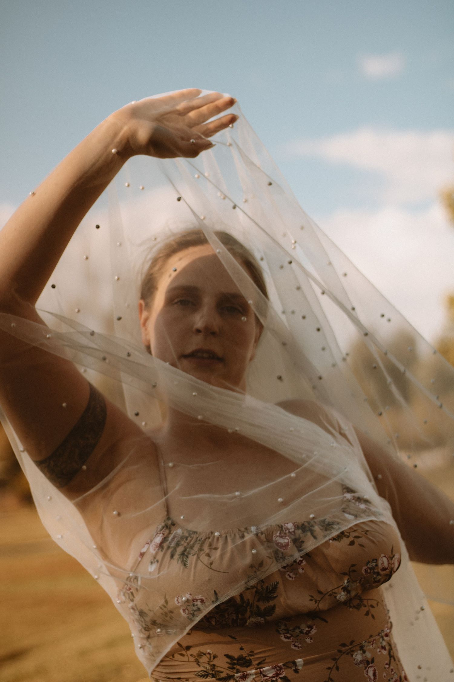 A figure in a floral dress stands in golden light with sheer fabric flowing in the wind against a cloudy sky.