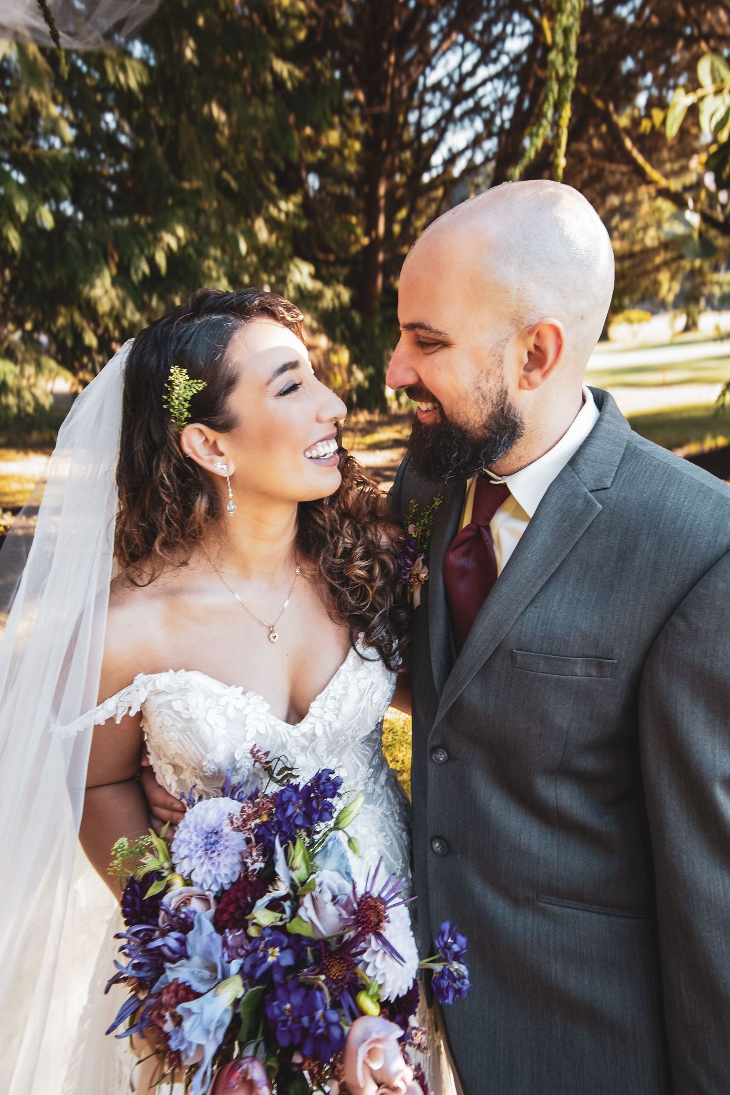 A joyful wedding couple shares a candid moment outdoors while the bride holds a stunning purple and blue bouquet.