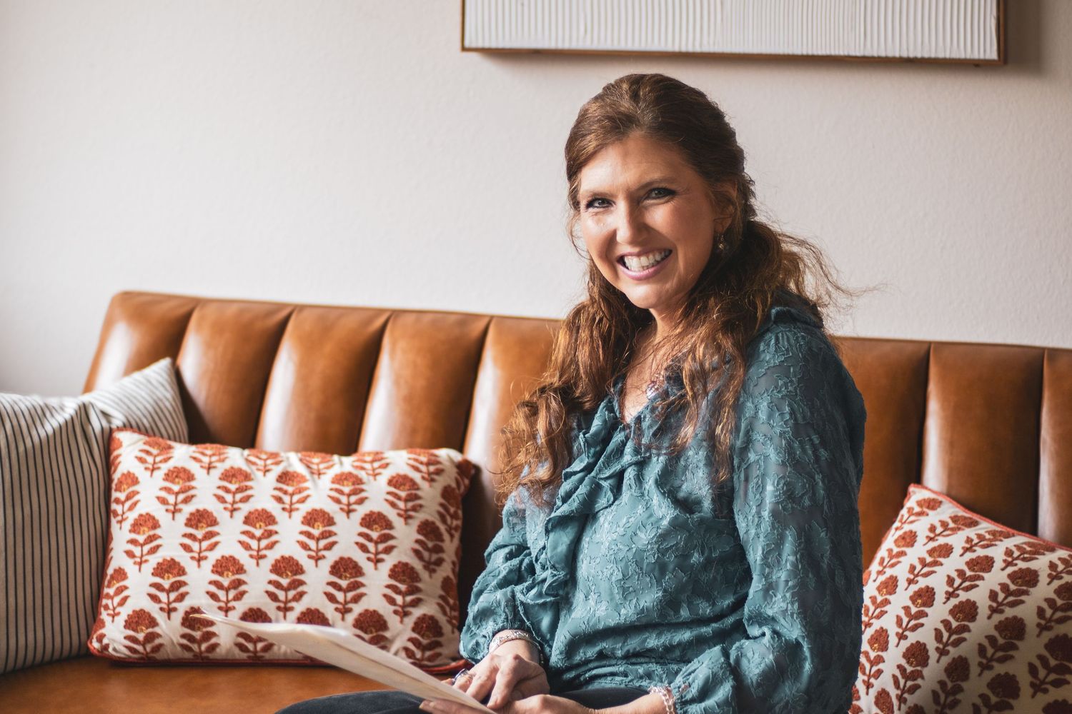 A person in a teal ruffled blouse sits comfortably on a brown leather couch with patterned throw pillows.