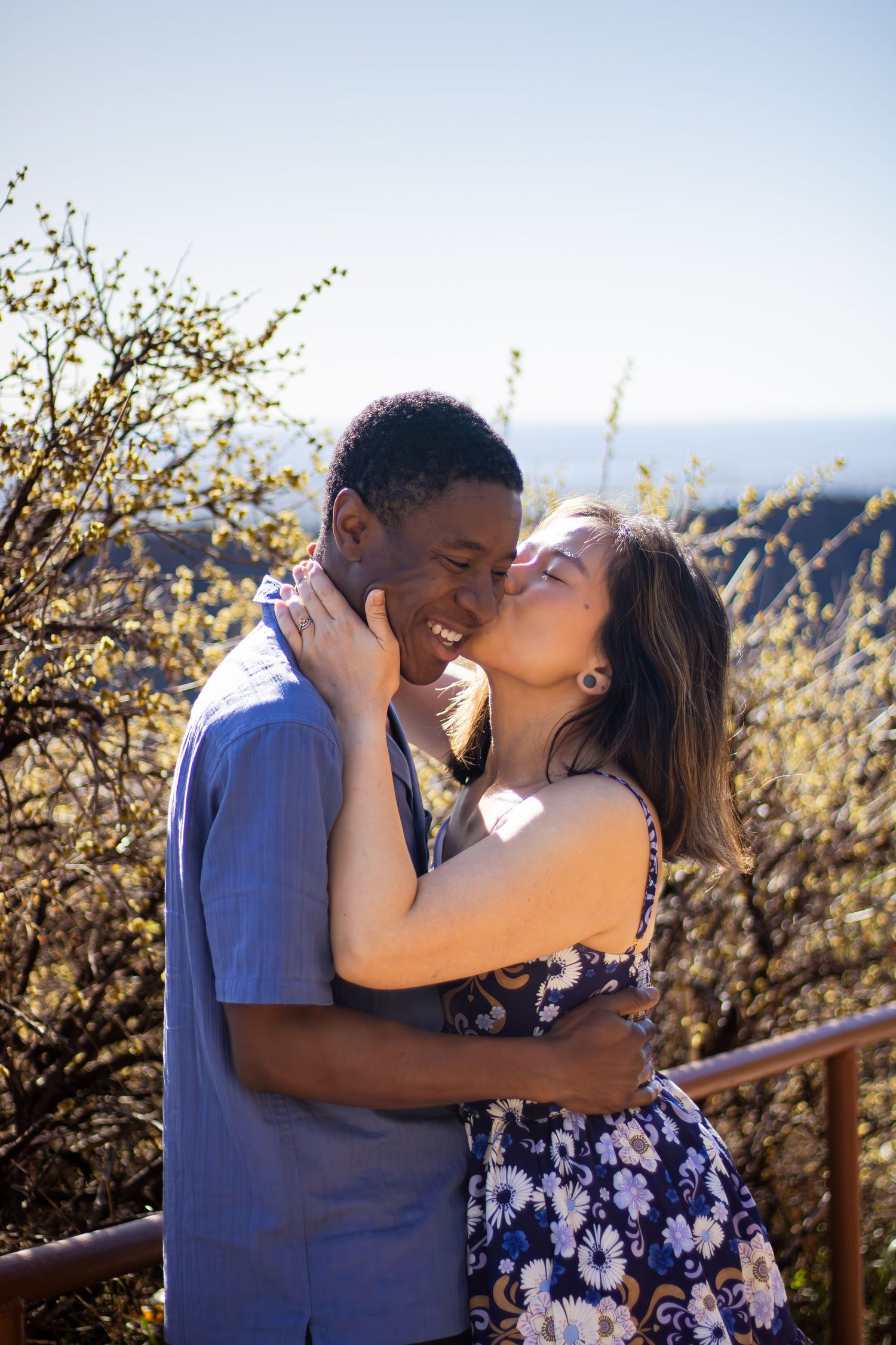 A couple shares a tender romantic moment outdoors on a sunny day with mountains and flowering bushes in the background.
