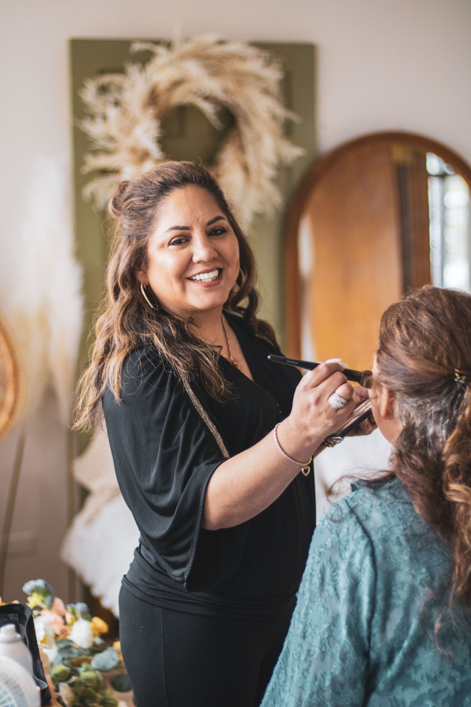 A makeup artist in black applies cosmetics to a client wearing a teal dress during a beauty session.