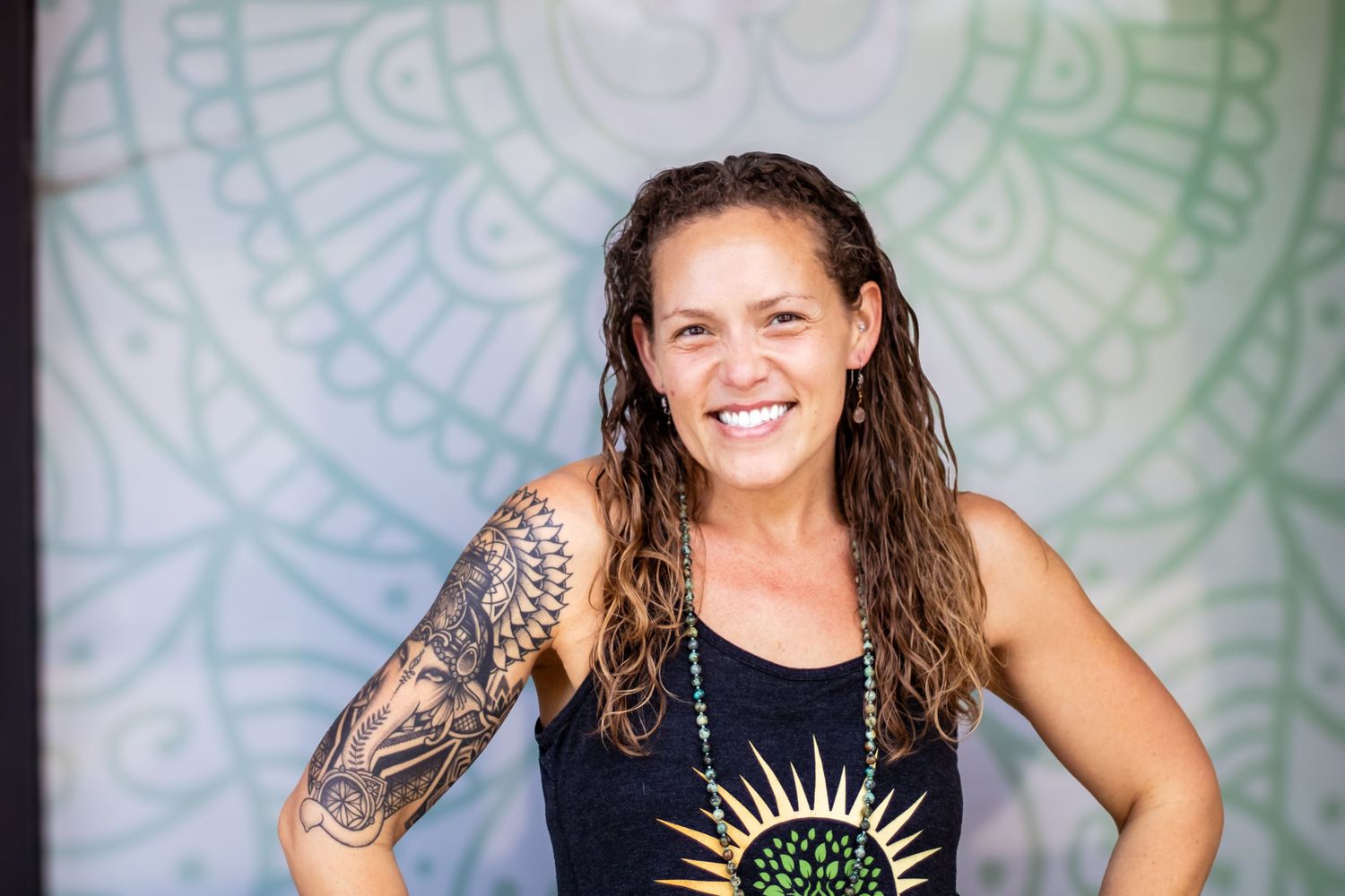 Athletic woman with wet hair and arm tattoo smiles while wearing a black tank top with sun design against patterned wall.