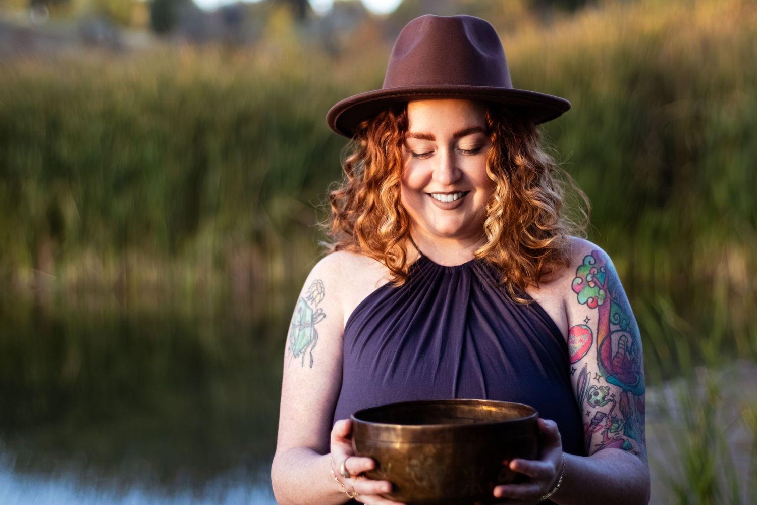 A person in a wide-brimmed hat holds a singing bowl in an outdoor setting with tall grass and natural light.