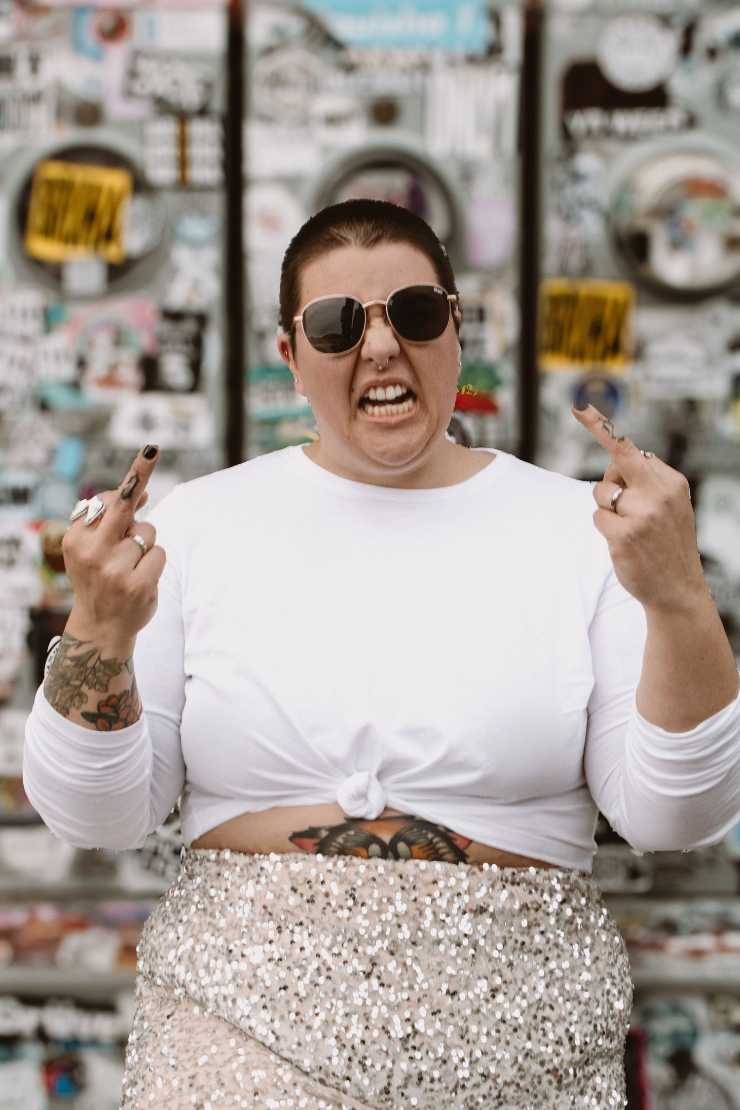 Person in white top with sunglasses making expressive hand gestures against a busy wall background.
