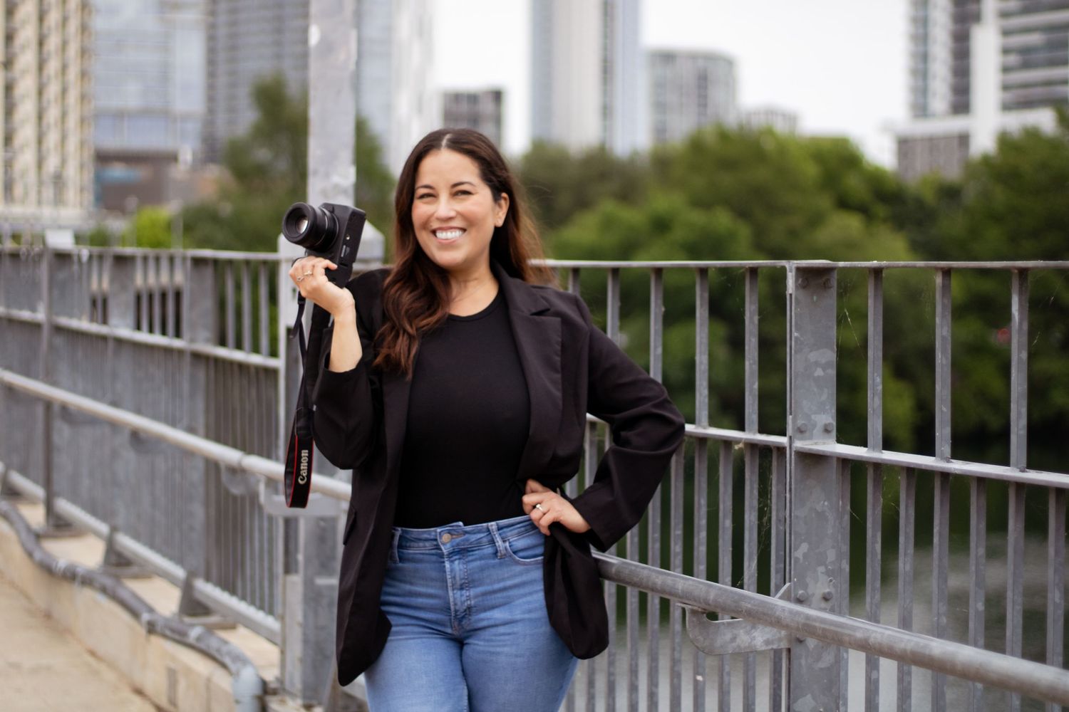 Photographer in black blazer and jeans holding camera on urban bridge with city skyline in background.