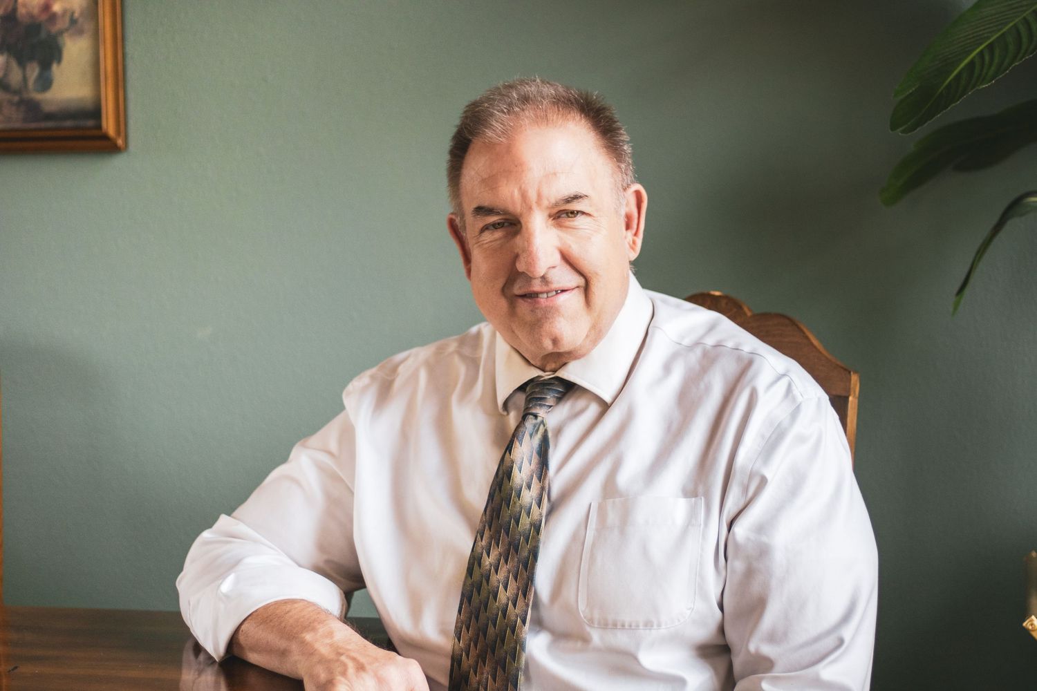 Professional portrait of someone in a white dress shirt and patterned tie seated at a desk in an office setting.