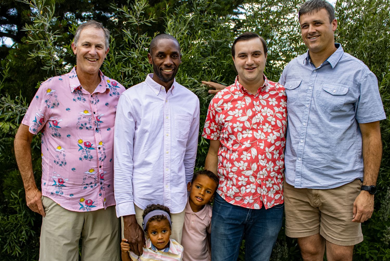 A group of people wearing colorful floral shirts and casual attire pose together outdoors on a sunny day.