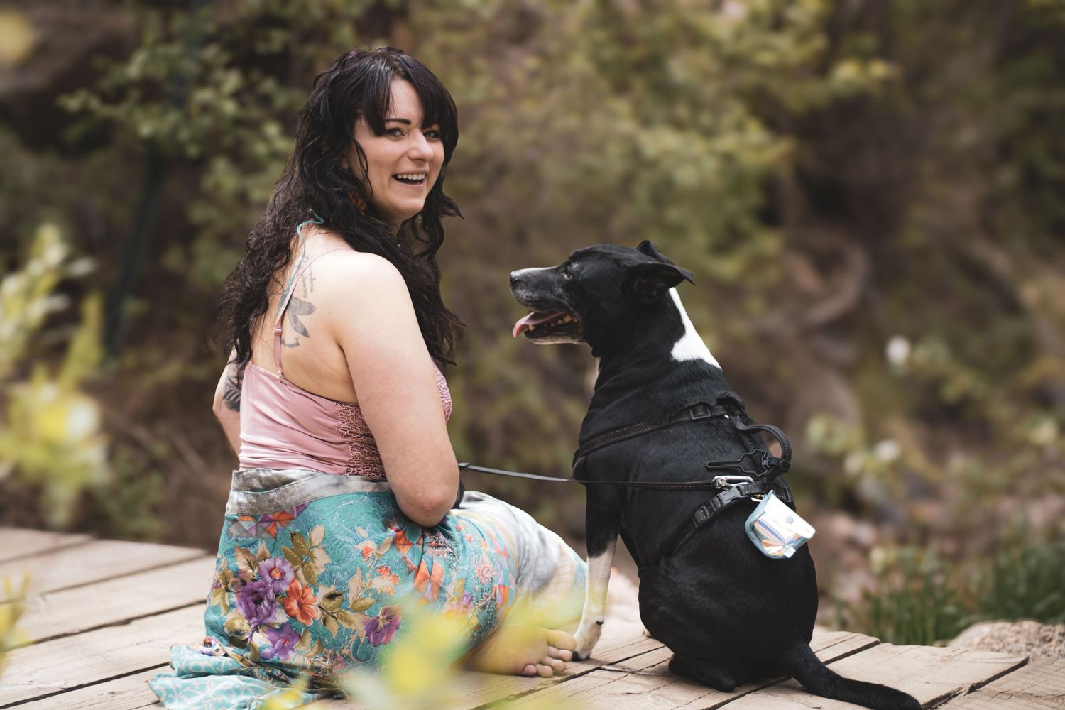 A person in a floral dress sits on a wooden deck railing while looking at their phone near a black backpack.