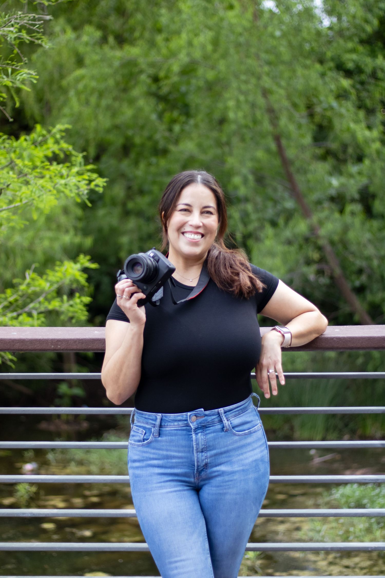 A photographer in a black top and jeans stands outdoors holding a camera against a green natural background.