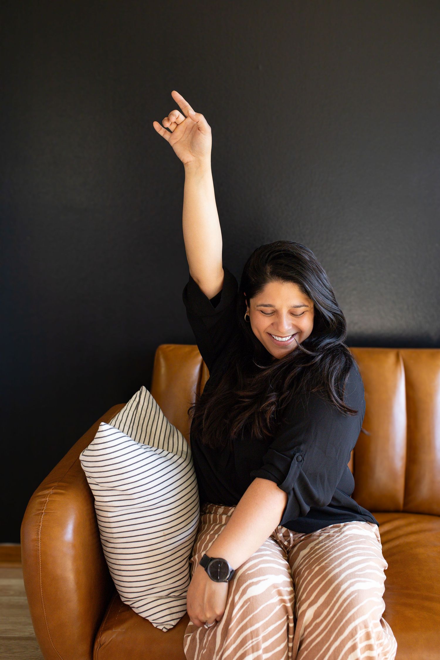 Woman in black top and beige pants sits on brown leather couch with striped pillow, raising arm joyfully.