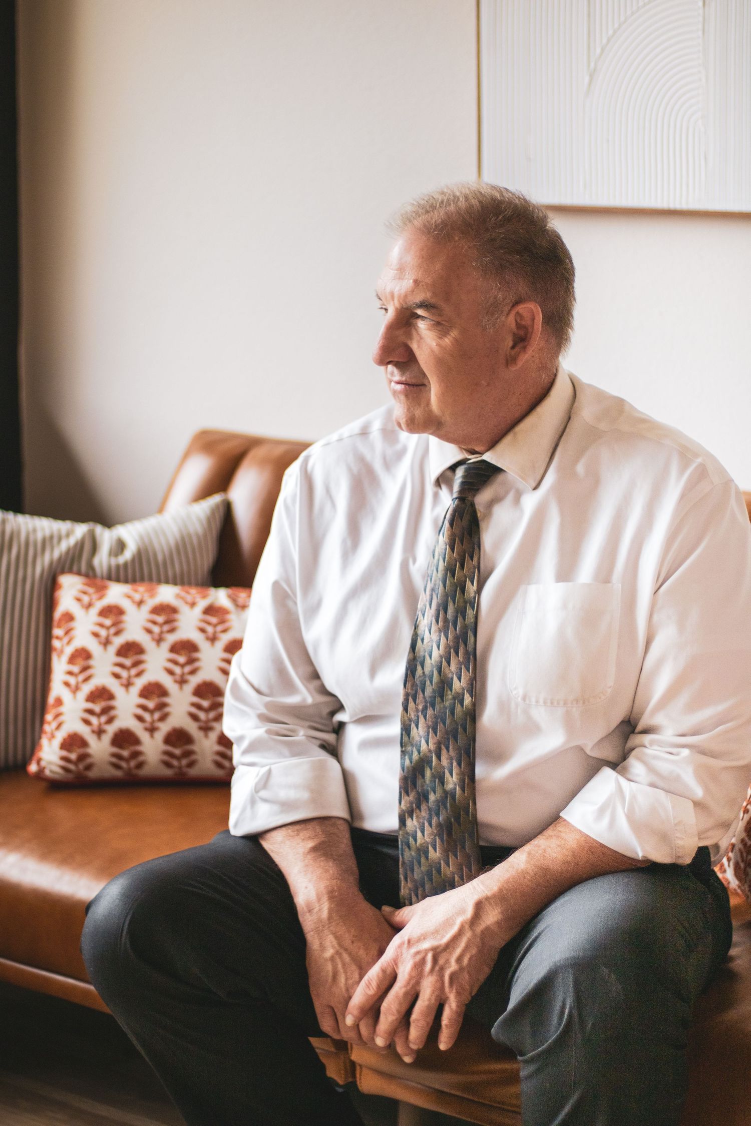 A person in a white dress shirt and tie sits contemplatively on a brown leather couch in a bright room.