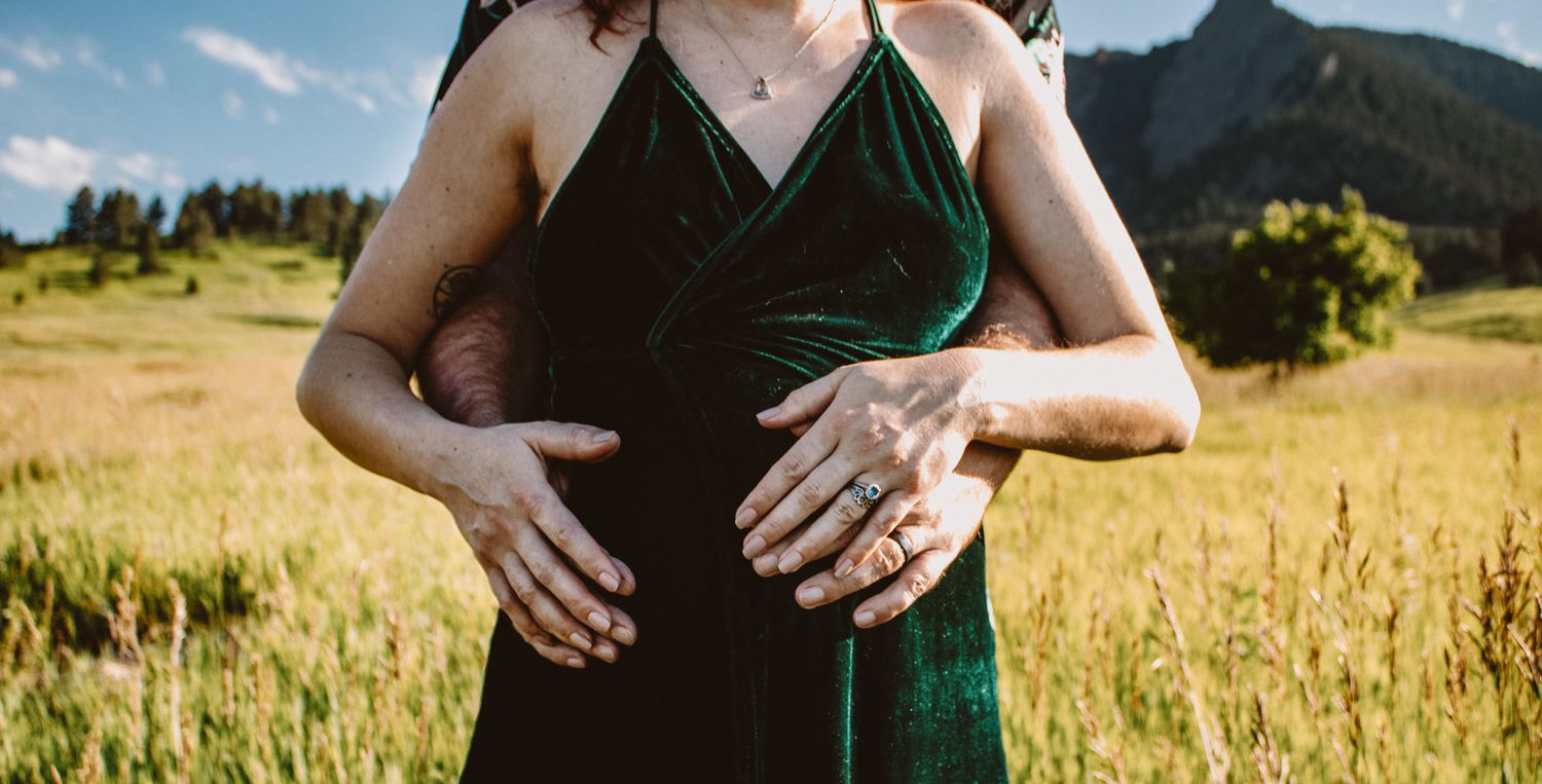 Two people embrace in a green dress and black outfit against a golden mountain meadow backdrop at sunset.