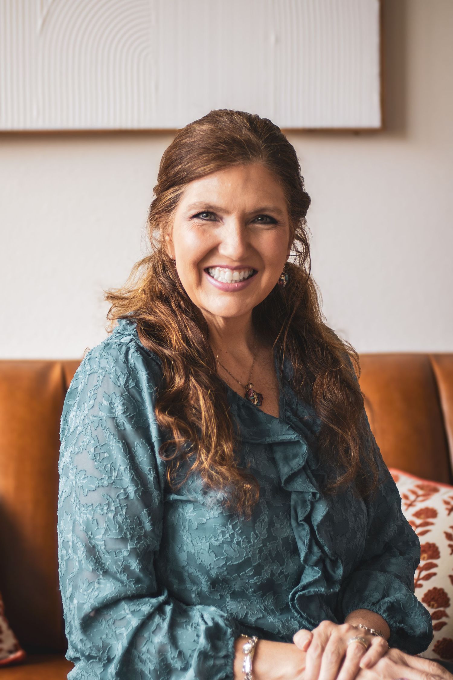 Woman in teal floral dress sits on brown leather couch and smiles warmly in bright natural light.