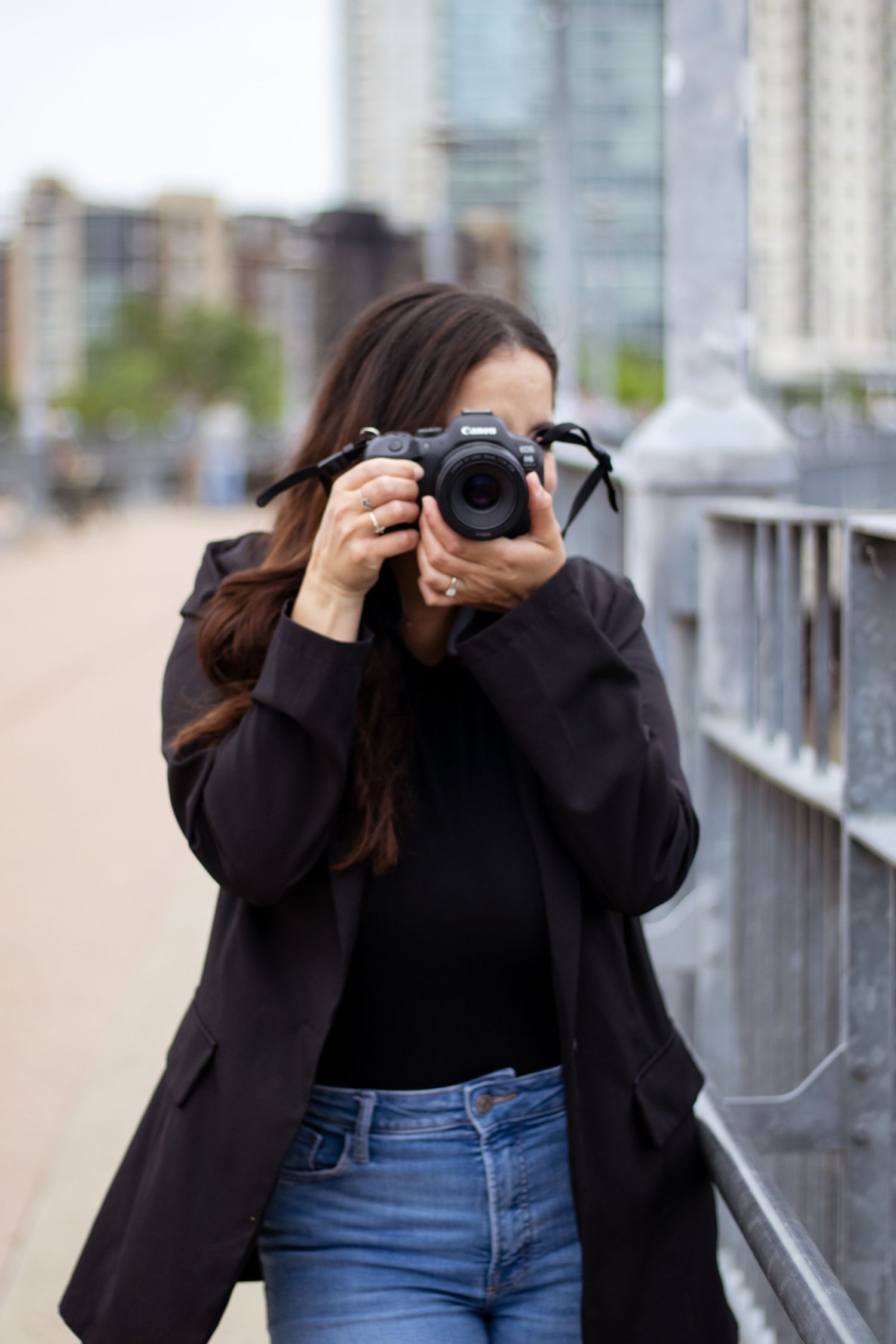 Photographer wearing black coat and jeans takes photo with professional camera on urban street.