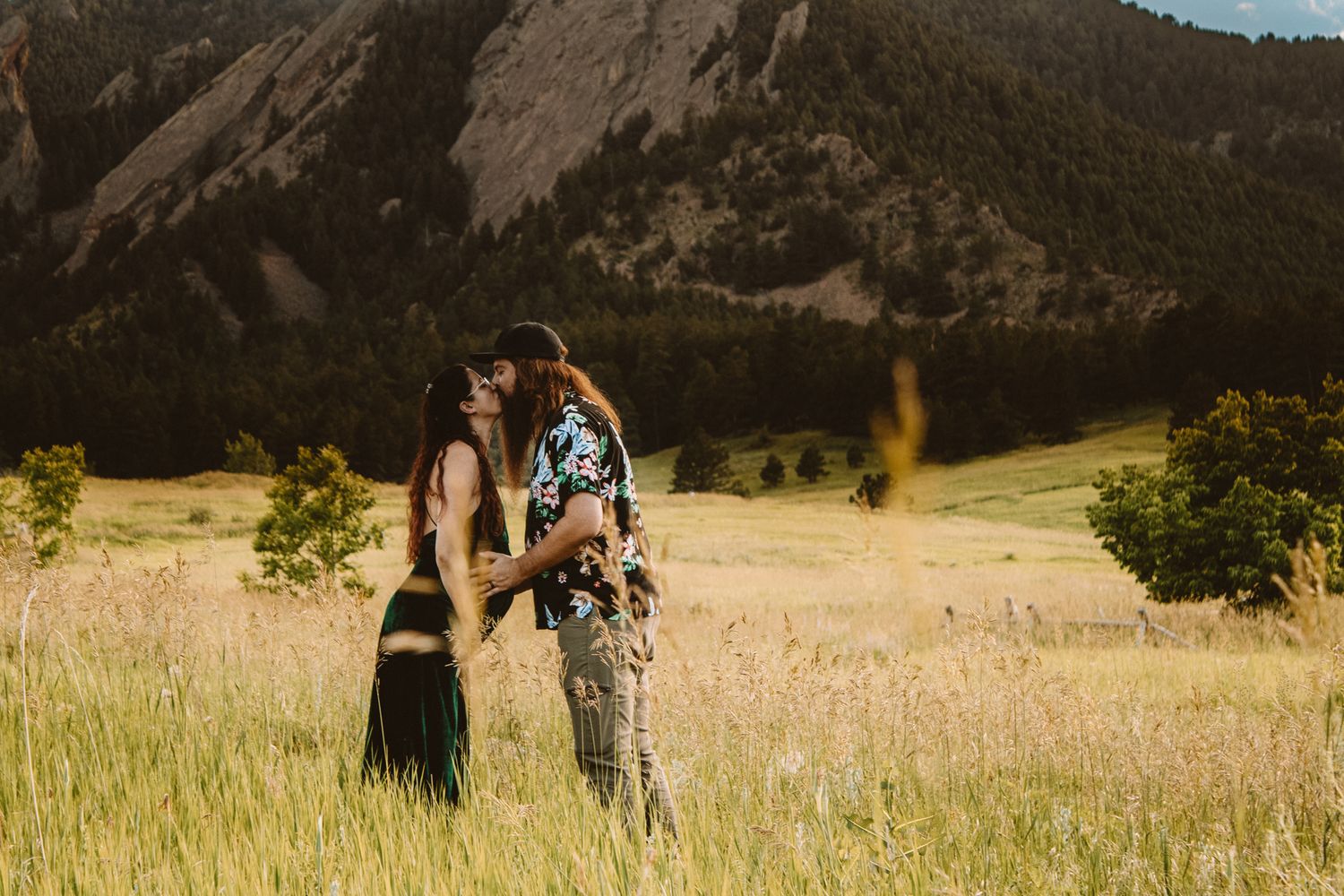 A couple in casual attire walks through a golden meadow with mountain views and sunlit scenery in the background.