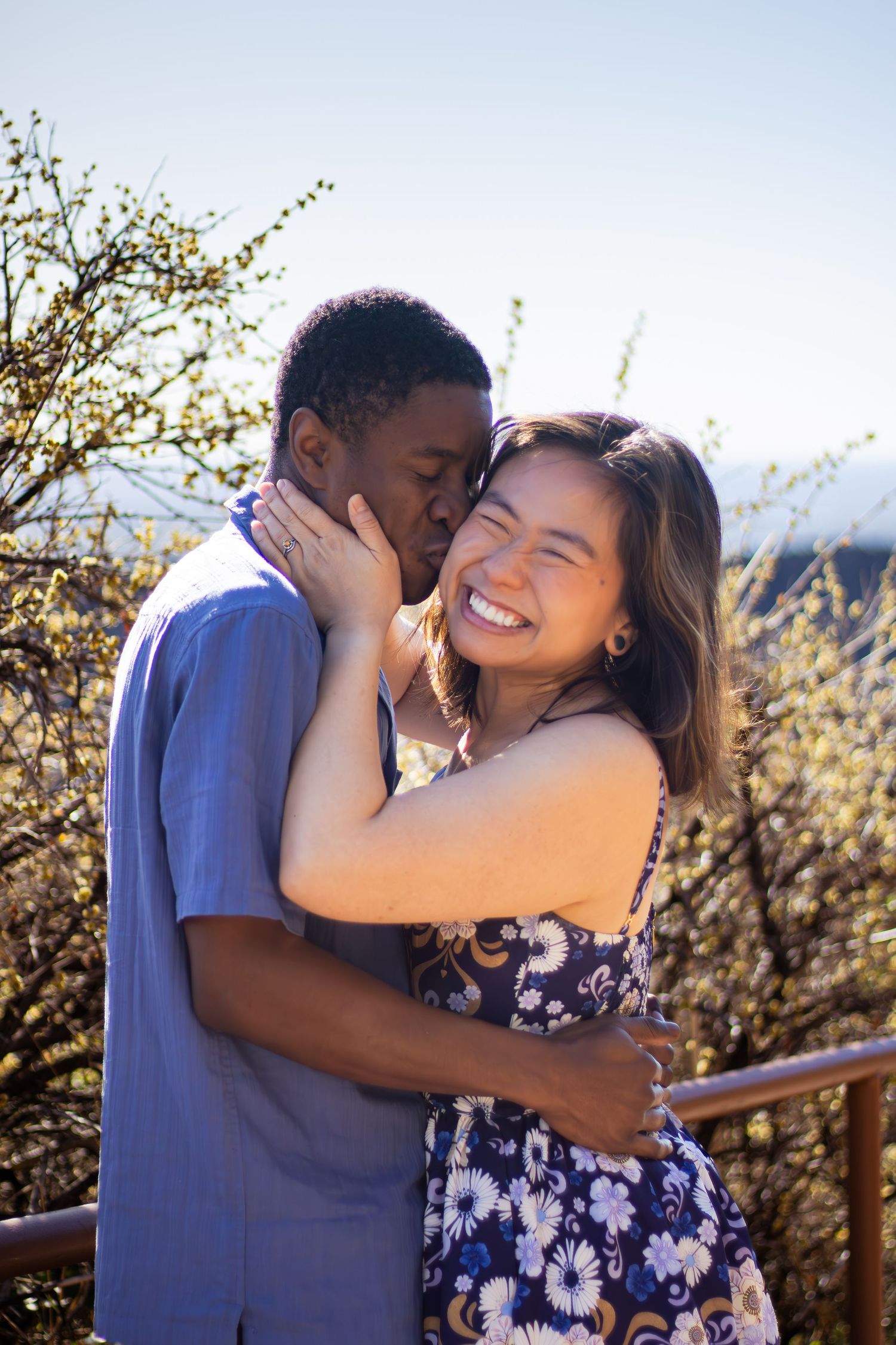 A joyful couple embraces outdoors on a sunny day with blooming trees in the background.