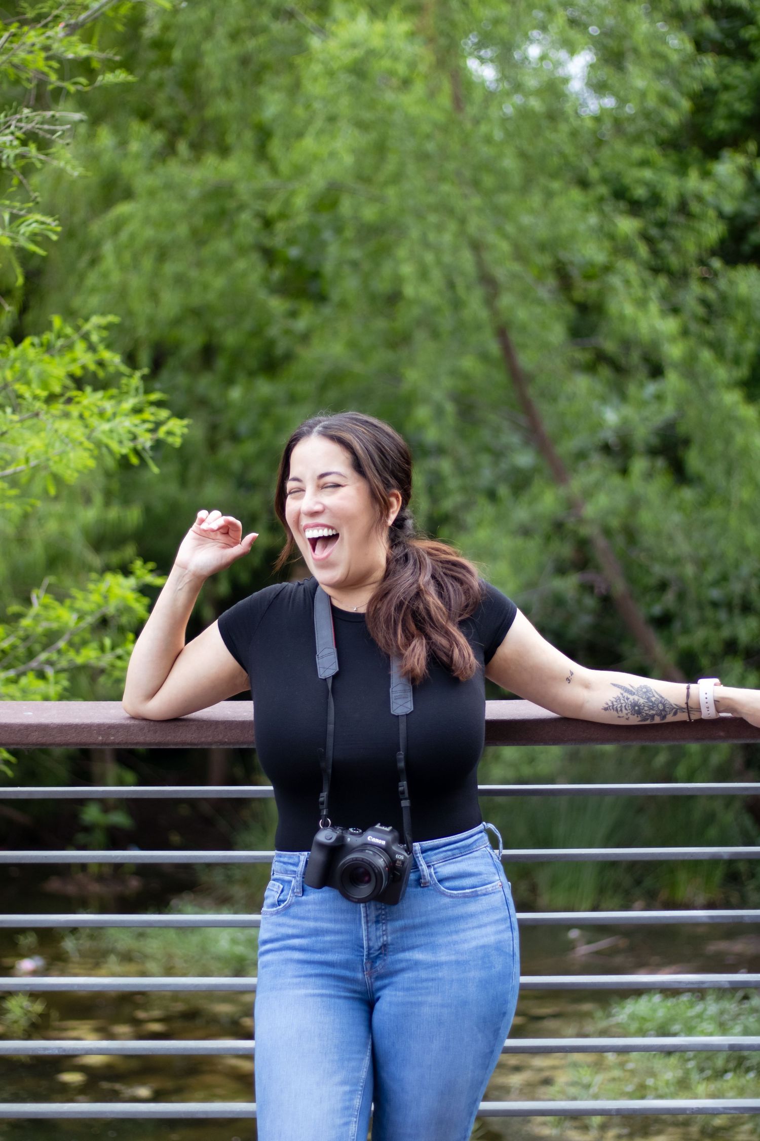 A photographer in a black top and jeans laughs while leaning on a railing outdoors with a camera around their neck.