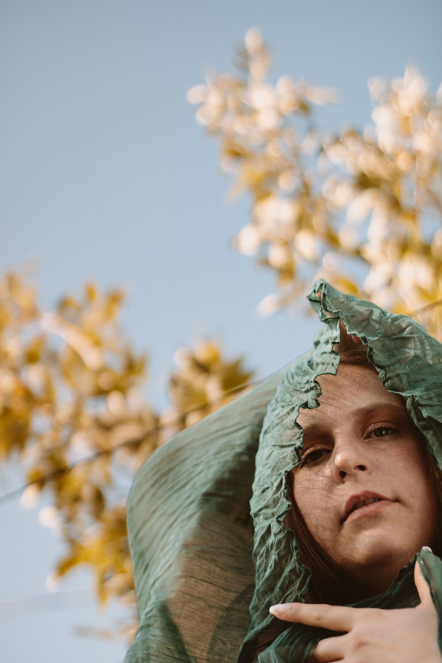 Artistic portrait with ruffled sage green fabric against autumn foliage and blue sky background.