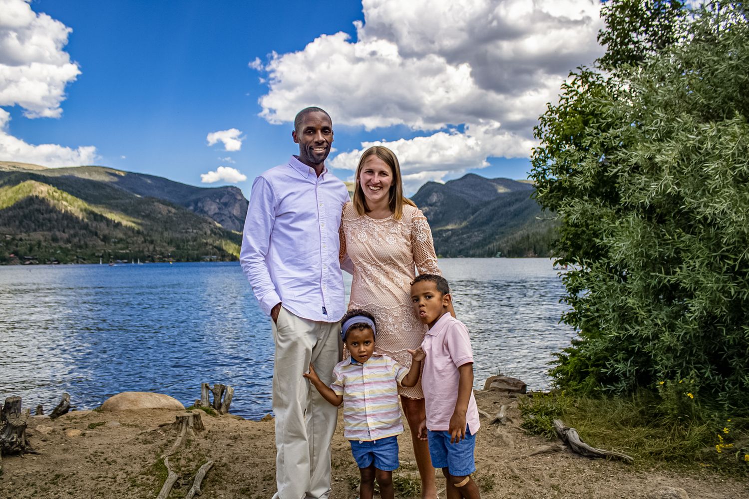 A scenic family photoshoot at a mountain lake with dramatic clouds and lush greenery in the background.