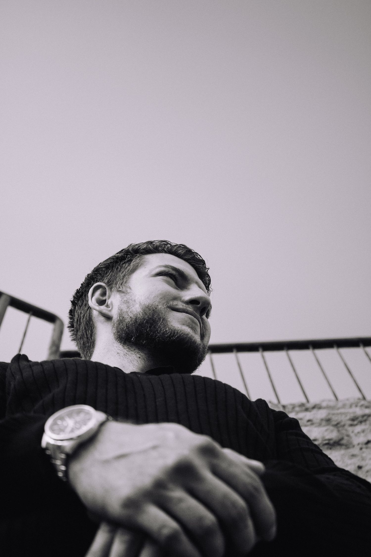 Low angle black and white portrait shot showing a person wearing a dark sweater with a watch visible on their wrist.