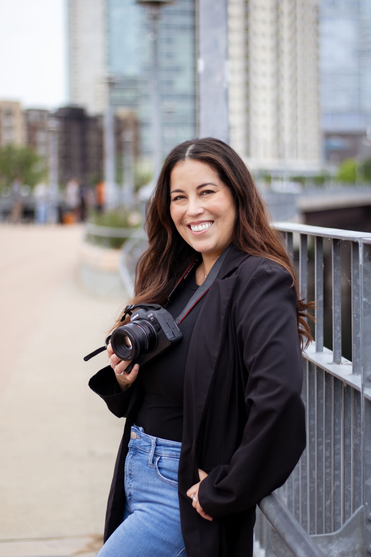 Photographer in a black blazer and jeans holds a camera while smiling on an urban walkway with city buildings in background.