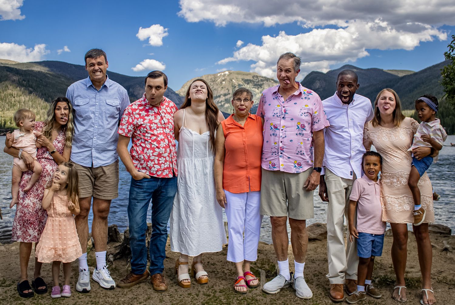 Large family group poses together outdoors with mountains and lake in background wearing coordinated pink and coral outfits.