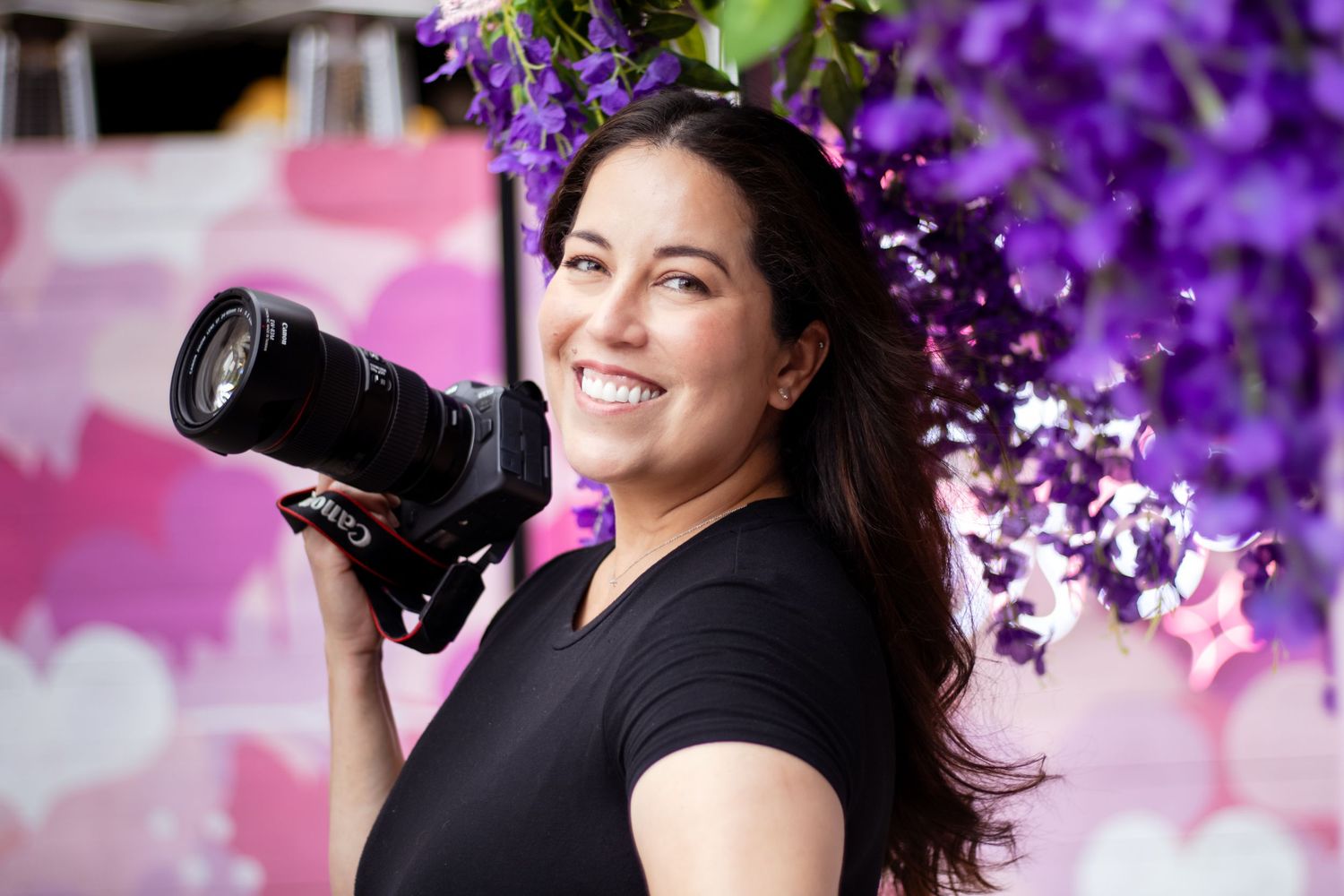 Professional photographer with long dark hair smiles while holding camera under blooming purple flowers in spring.