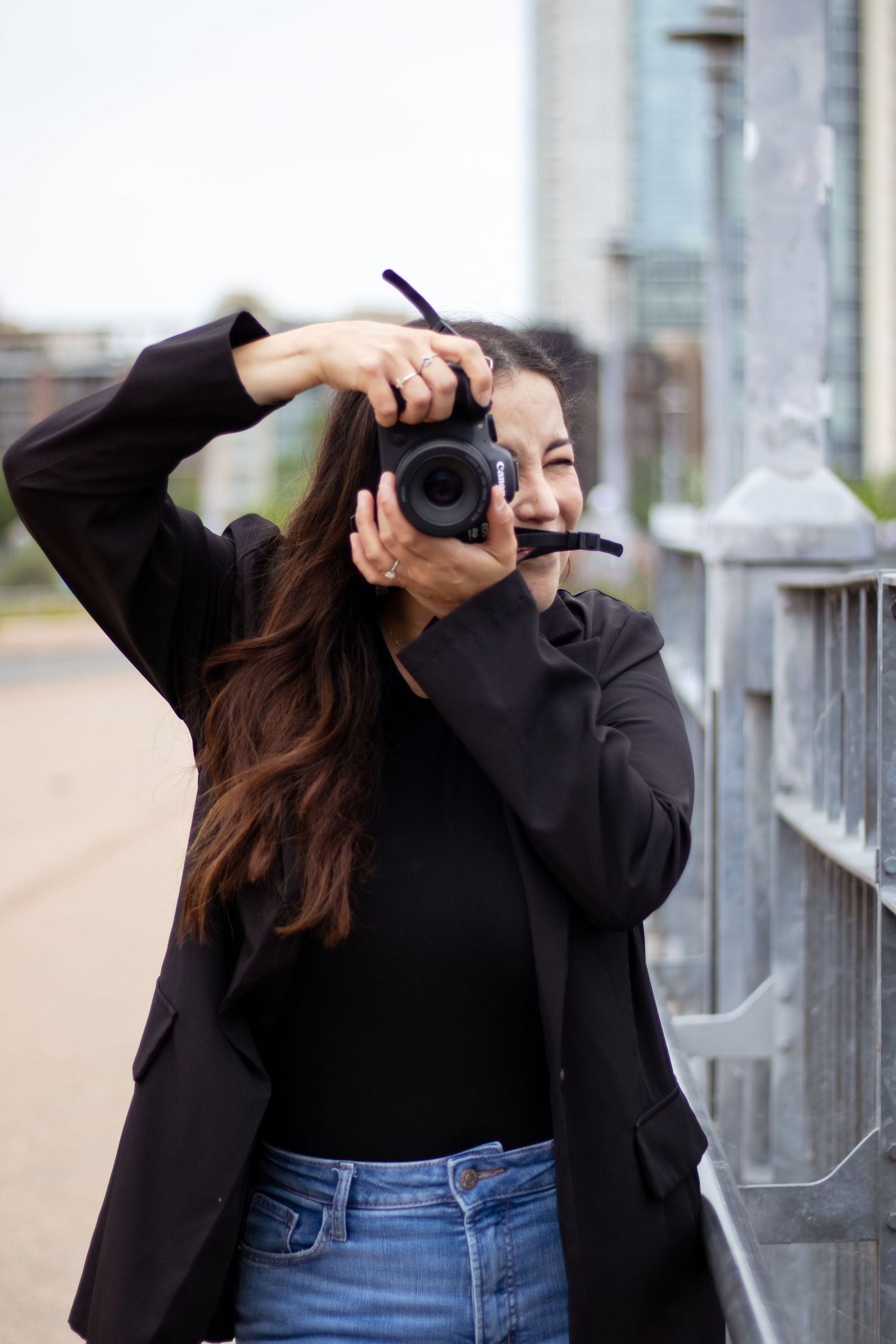 A photographer in a black jacket and jeans takes pictures with a professional camera along a railing outdoors.