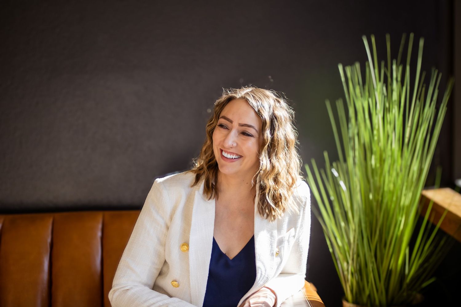 A person in a white blazer and navy top sits on a brown leather bench with a green plant in the background.