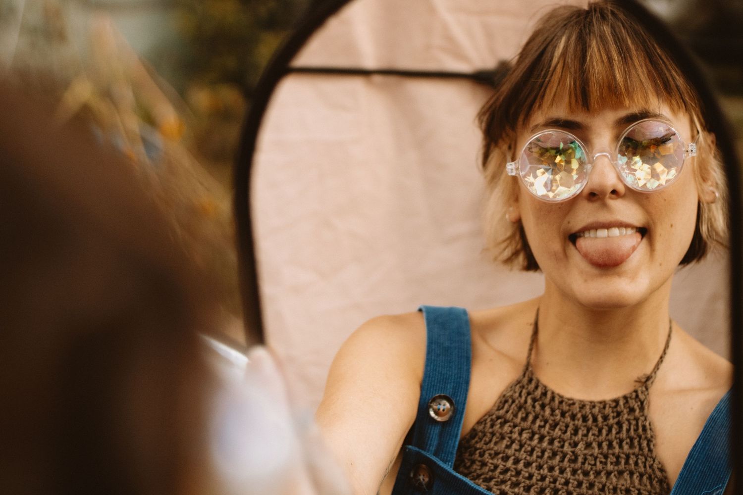Someone wearing round glasses decorated with daisy flowers sticks out their tongue playfully in a sunny outdoor setting.
