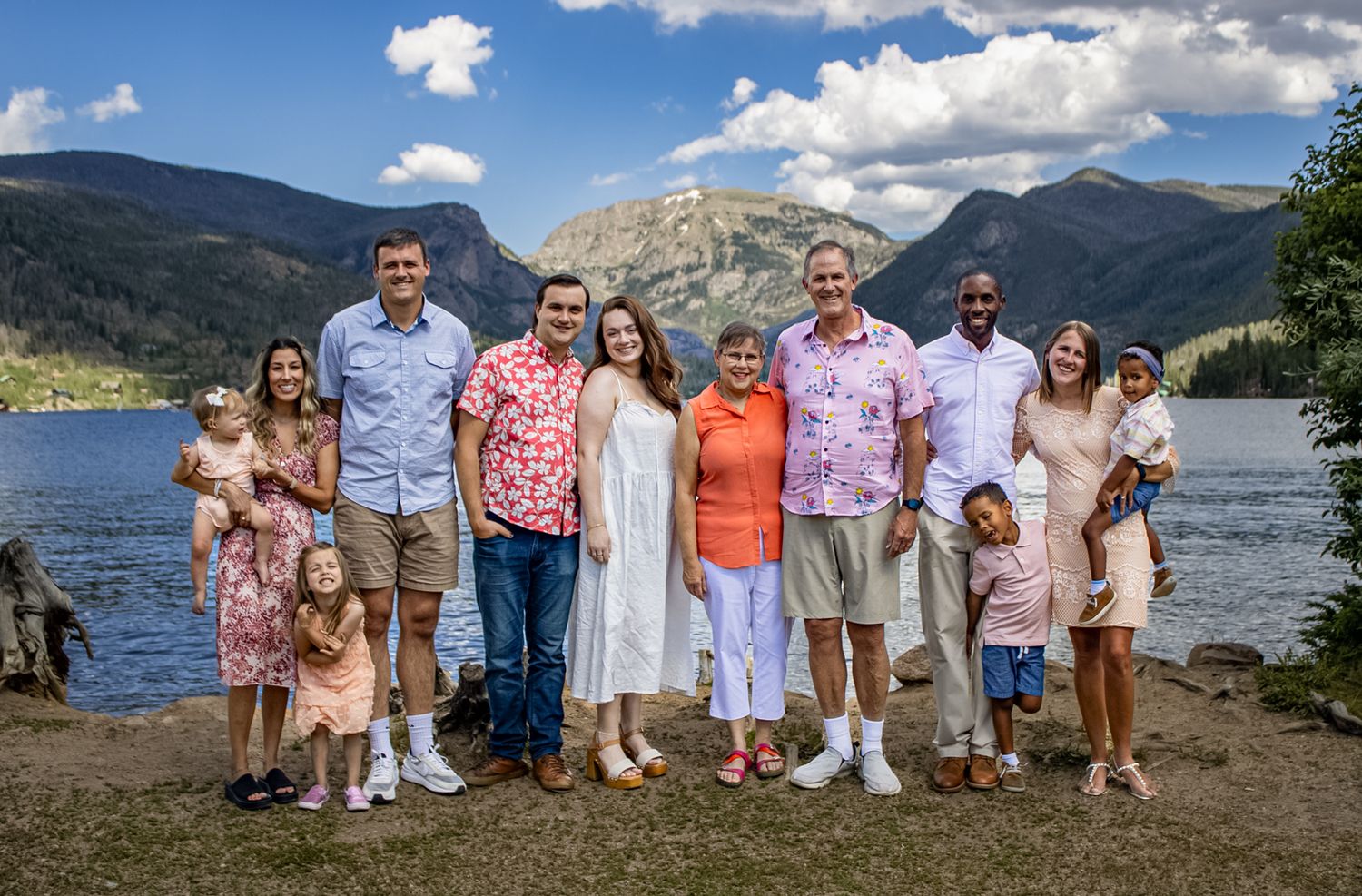 Group of people gathered outdoors against a scenic mountain lake backdrop with blue skies and clouds.