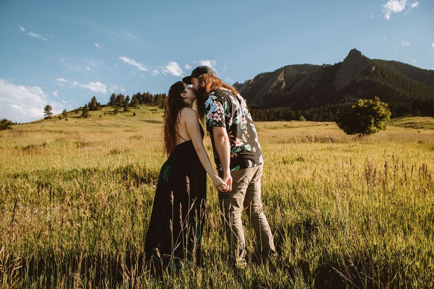Couple embraces in golden mountain meadow during sunset with dramatic mountain peaks in background.
