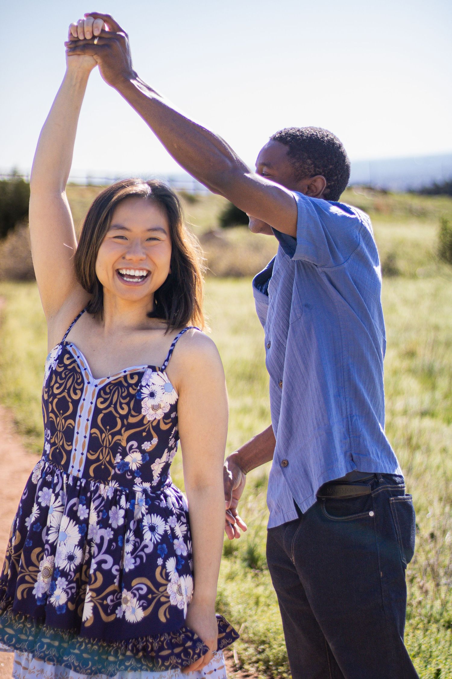 A couple dances playfully together in a sunny meadow, with the woman wearing a floral sundress.