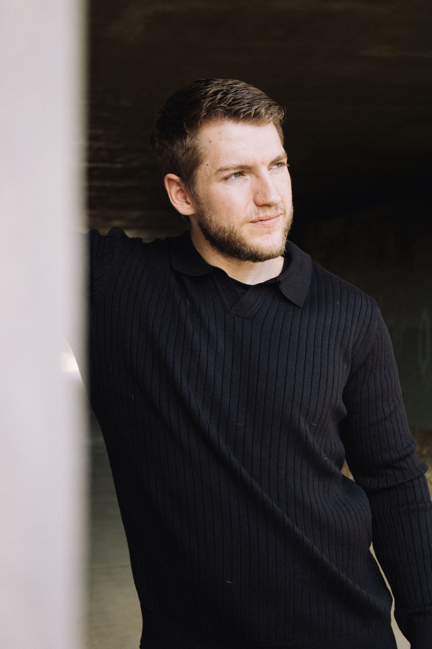 Portrait photo through white columns showing someone wearing a black ribbed sweater looking to the side.