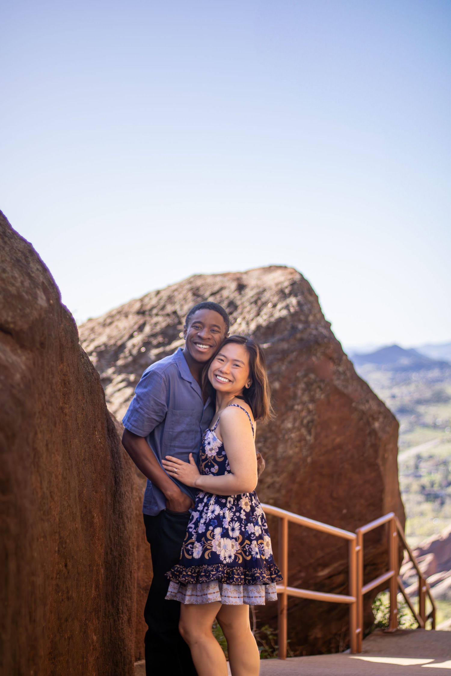 A happy couple embraces while standing on a scenic mountain overlook with rocky terrain and blue skies in the background.