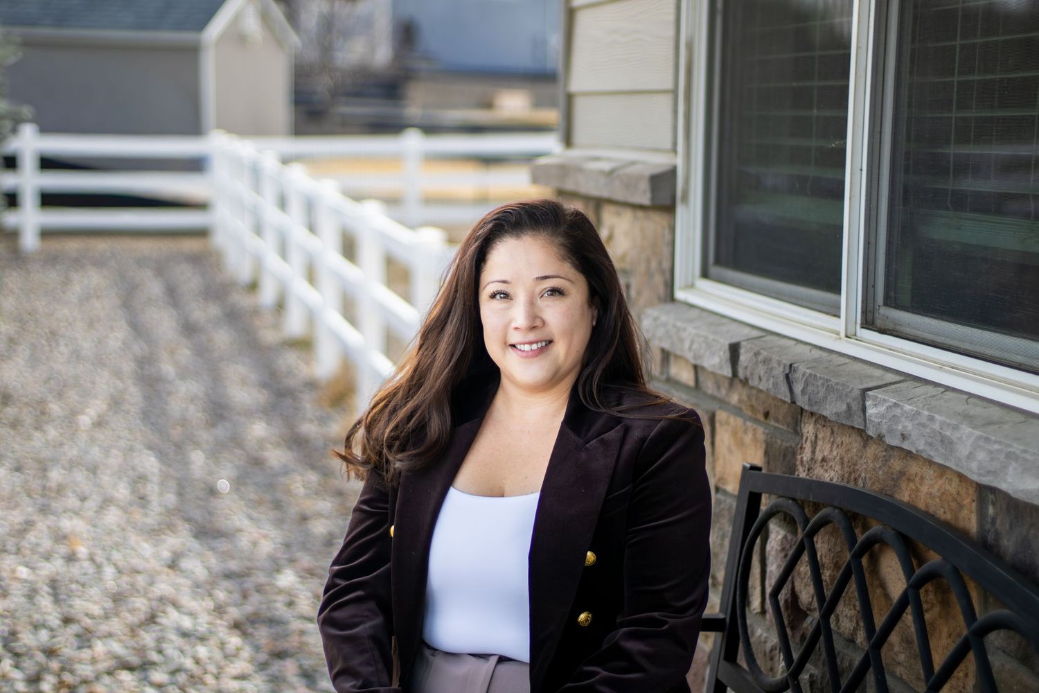 A person in a black blazer and white top sits on a porch with white fencing and stone accents in the background.