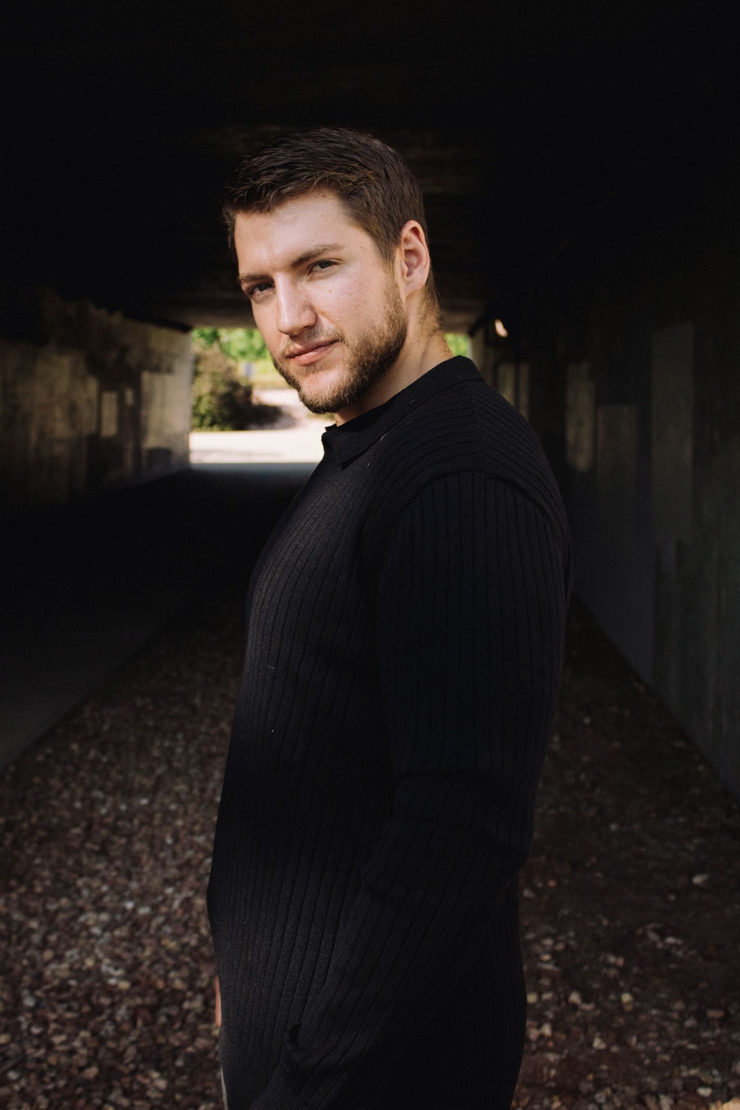Portrait of person wearing black sweater standing in dark tunnel with natural lighting in background.