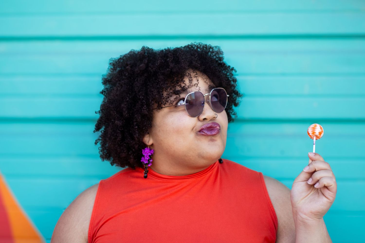 Person in red shirt poses playfully against turquoise wall while holding a lollipop.